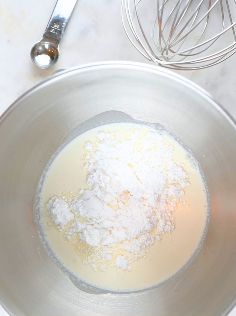 Ingredients for whipped cream with vanilla in a mixing bowl, shown next to a measuring spoon and wire whisk attachment