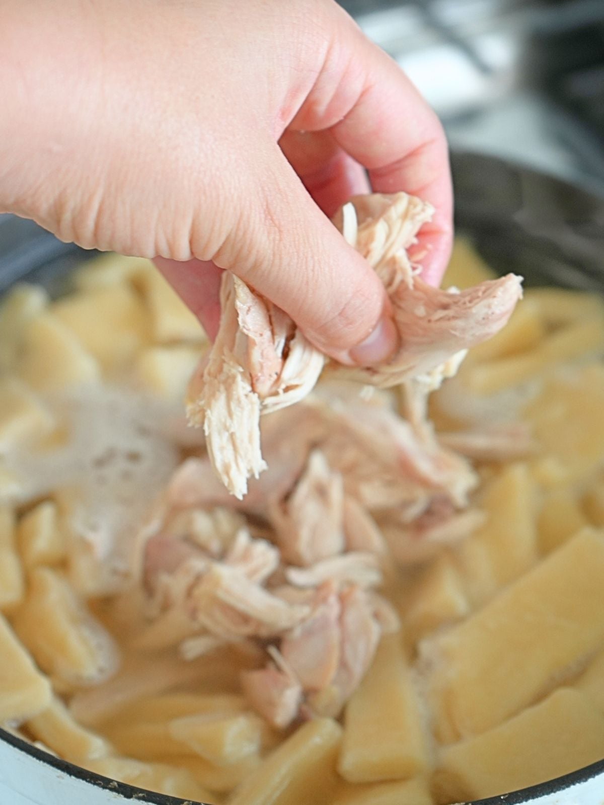A hand placing shredded chicken in a pot of broth and pastry.