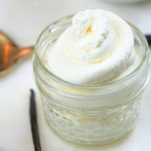 A small jar of homemade vanilla whipped cream with a spoon and vanilla beans in the background