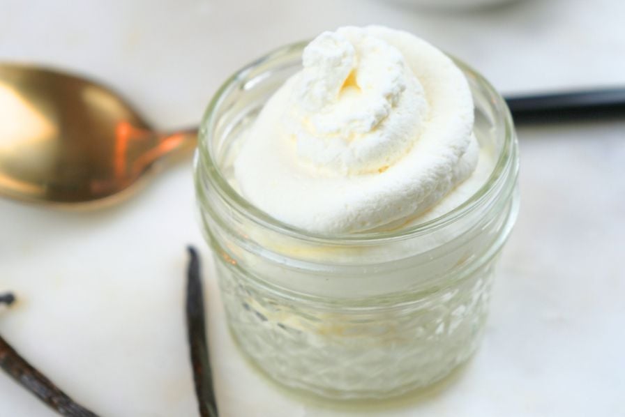 A small jar of homemade vanilla whipped cream with a spoon and vanilla beans in the background