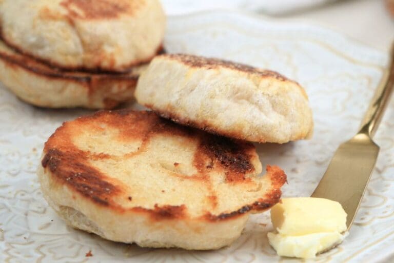 A toasted English muffin on a decorative plate next to a gold knife with butter
