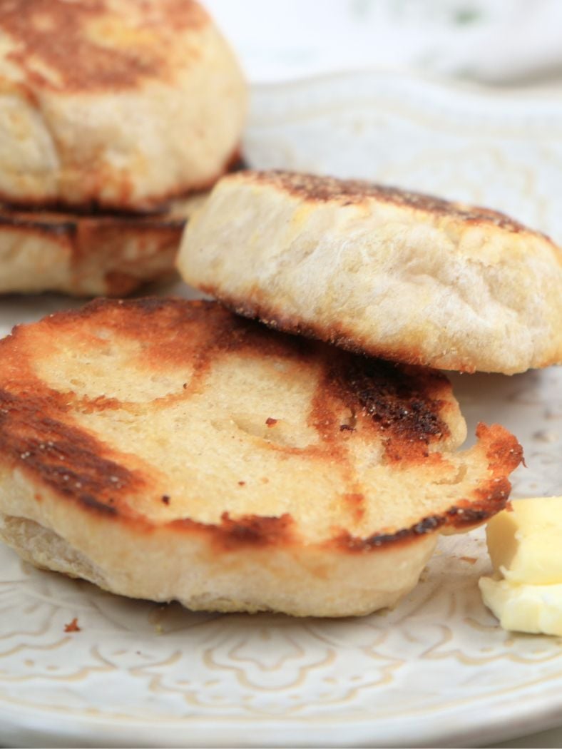 A toasted sourdough English muffin on a plate next to a knife with butter on it