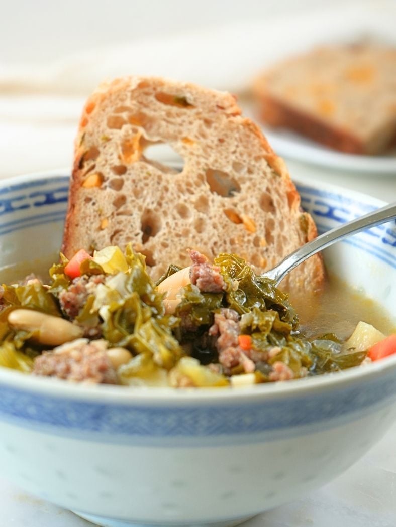 A bowl of sausage, kale, and white bean soup with a slice of sourdough bread in it