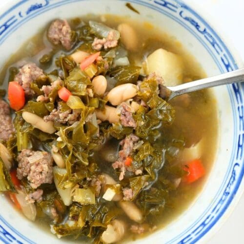 An close up of a bowl of sausage, kale, and white bean soup in a blue bowl with a spoon in it