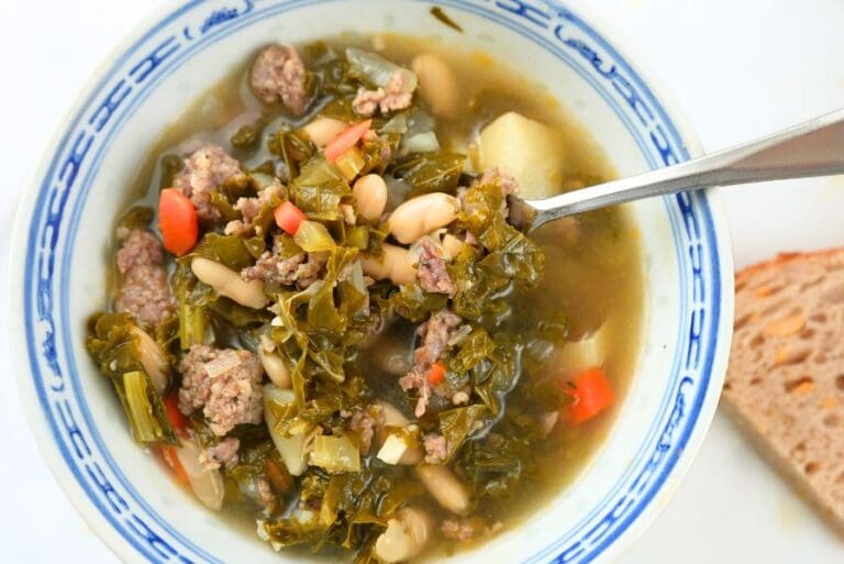 An close up of a bowl of sausage, kale, and white bean soup in a blue bowl with a spoon in it