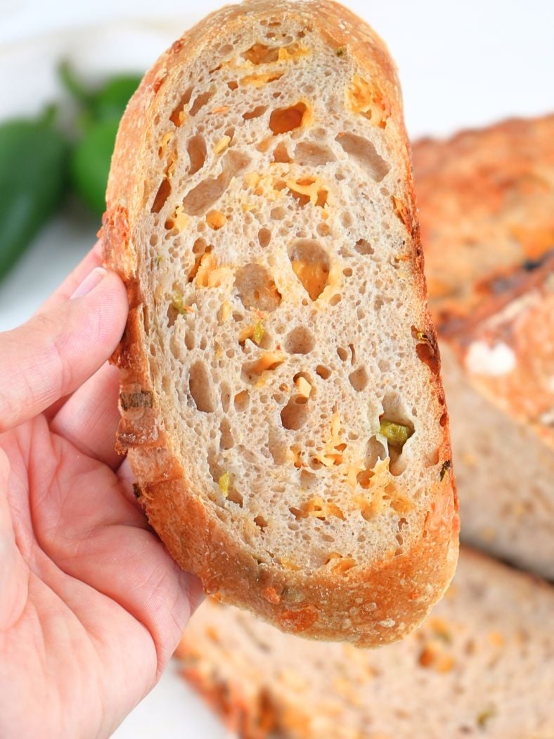 An hand holding a slice of sourdough cheddar jalapeño bread with bread and peppers in the background