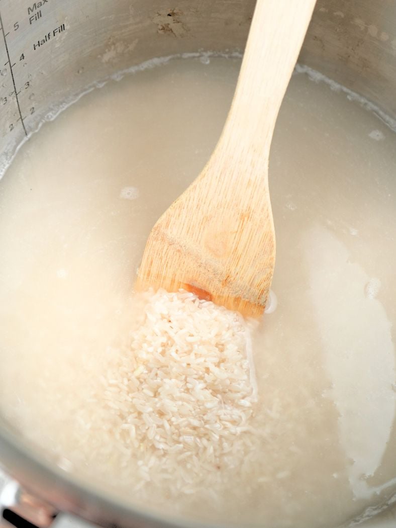 Water and rice mixed together in an Instant Pot, shown with a wooden spoon