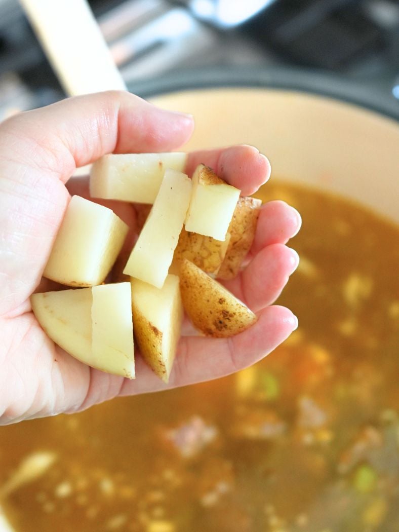 A hand holding chopped potatoes over a pot of soup