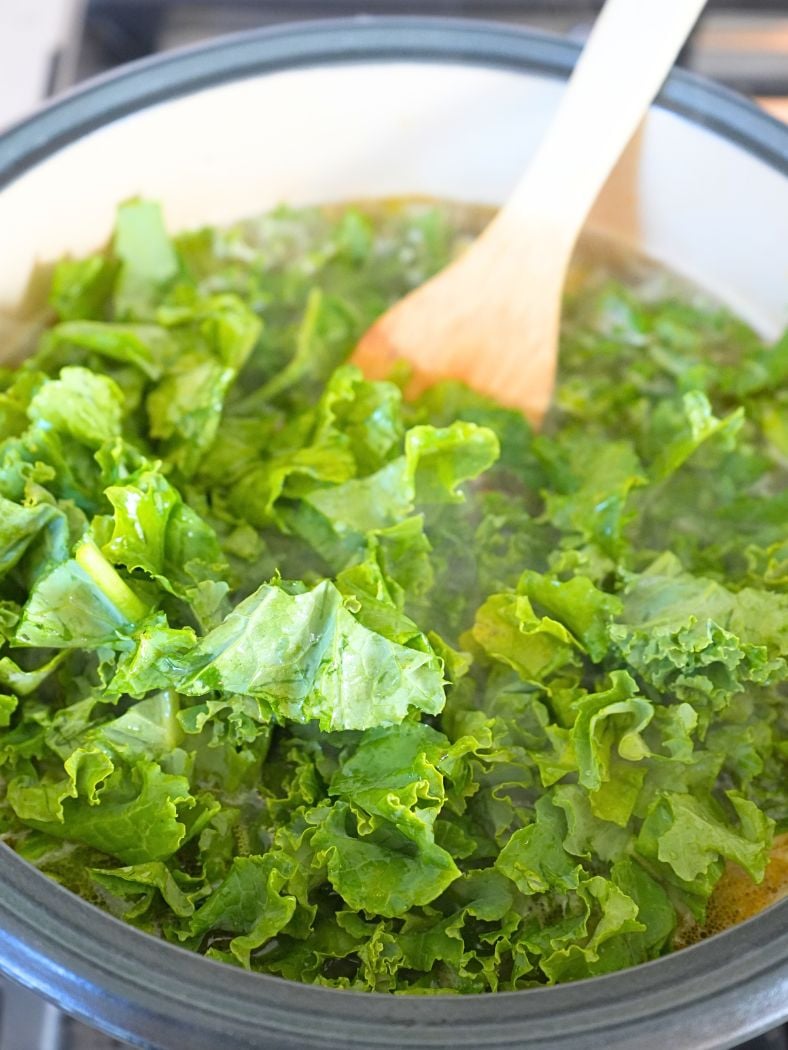 Chopped kale in a Dutch oven, being stirred into soup