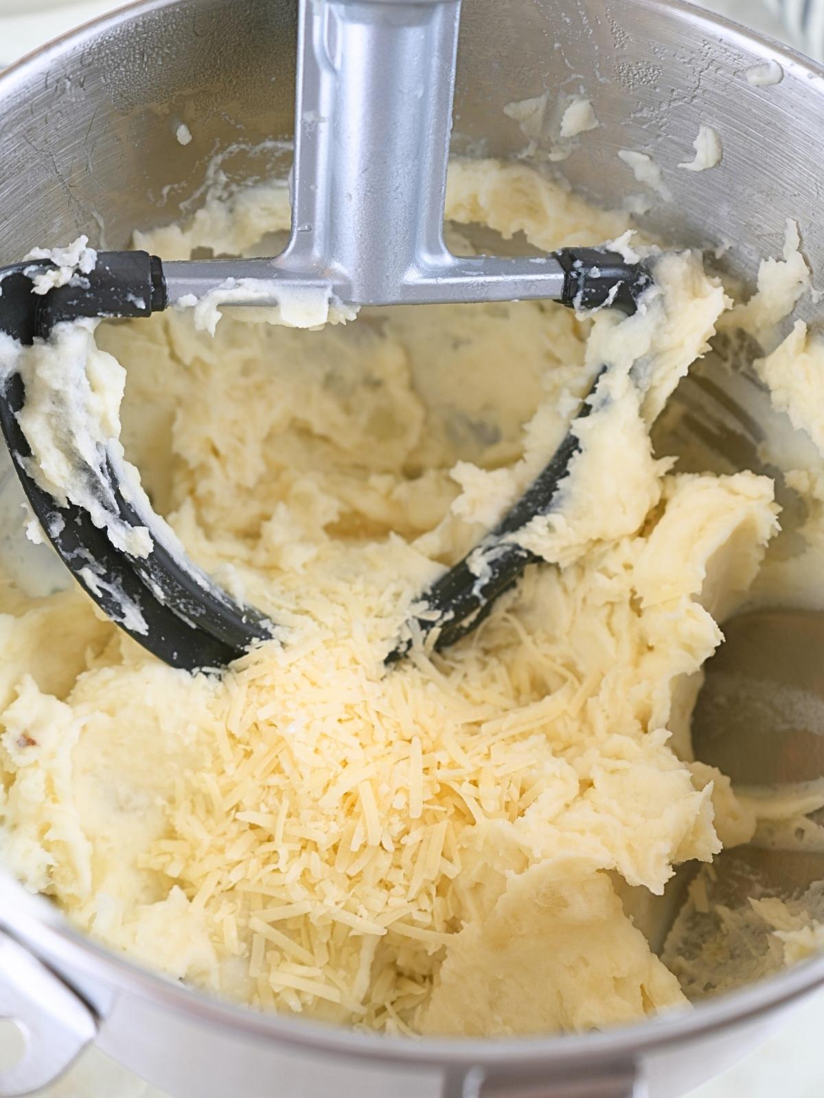 Mashed potatoes in a bowl with shredded parmesan cheese, shown with a paddle attachment.