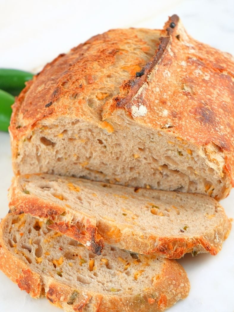 A boule of sourdough cheddar jalapeño bread, sliced and with fresh peppers in the background