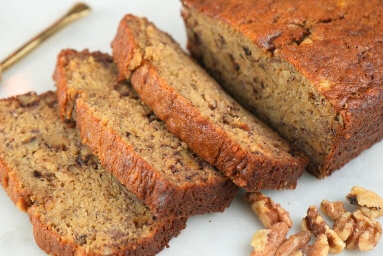 A loaf of sourdough discard banana bread sliced and next to walnuts and a gold knife