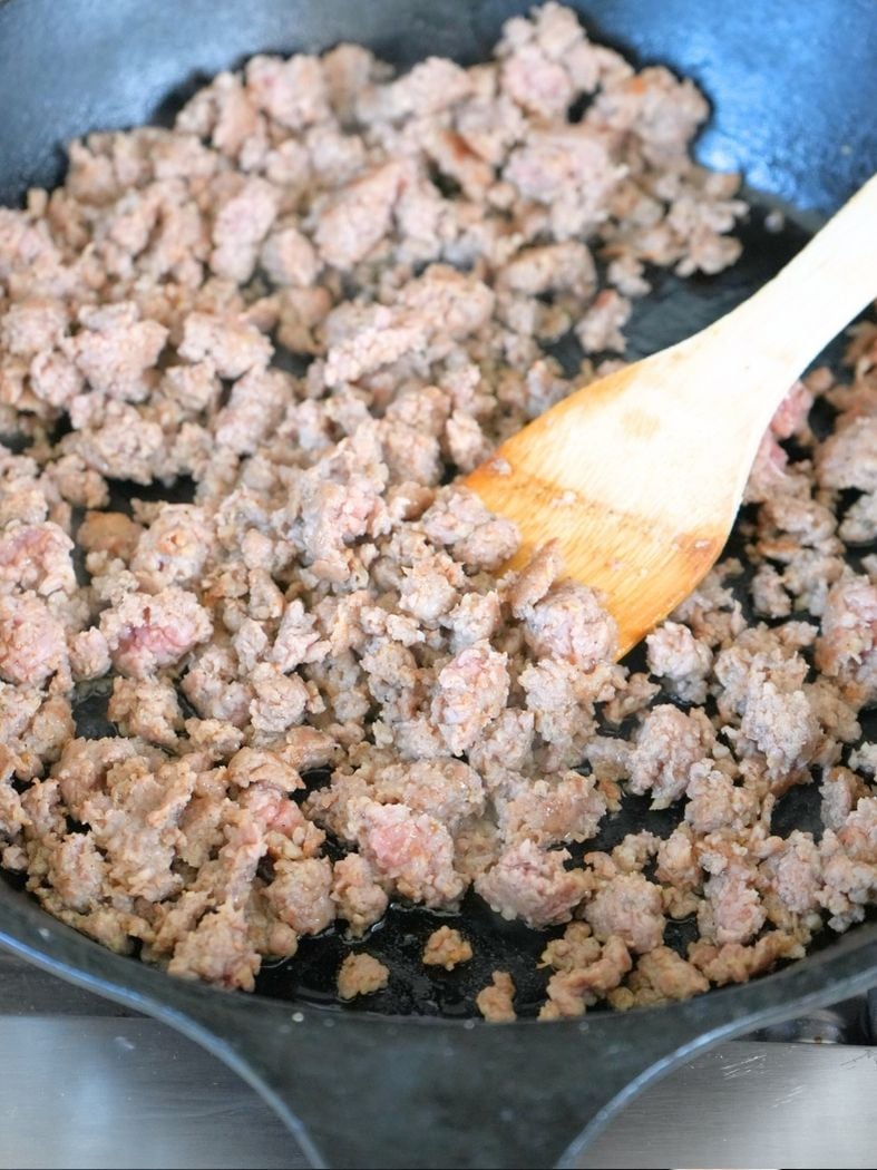 Ground sausage browning in a cast iron skillet, shown with wooden spatula
