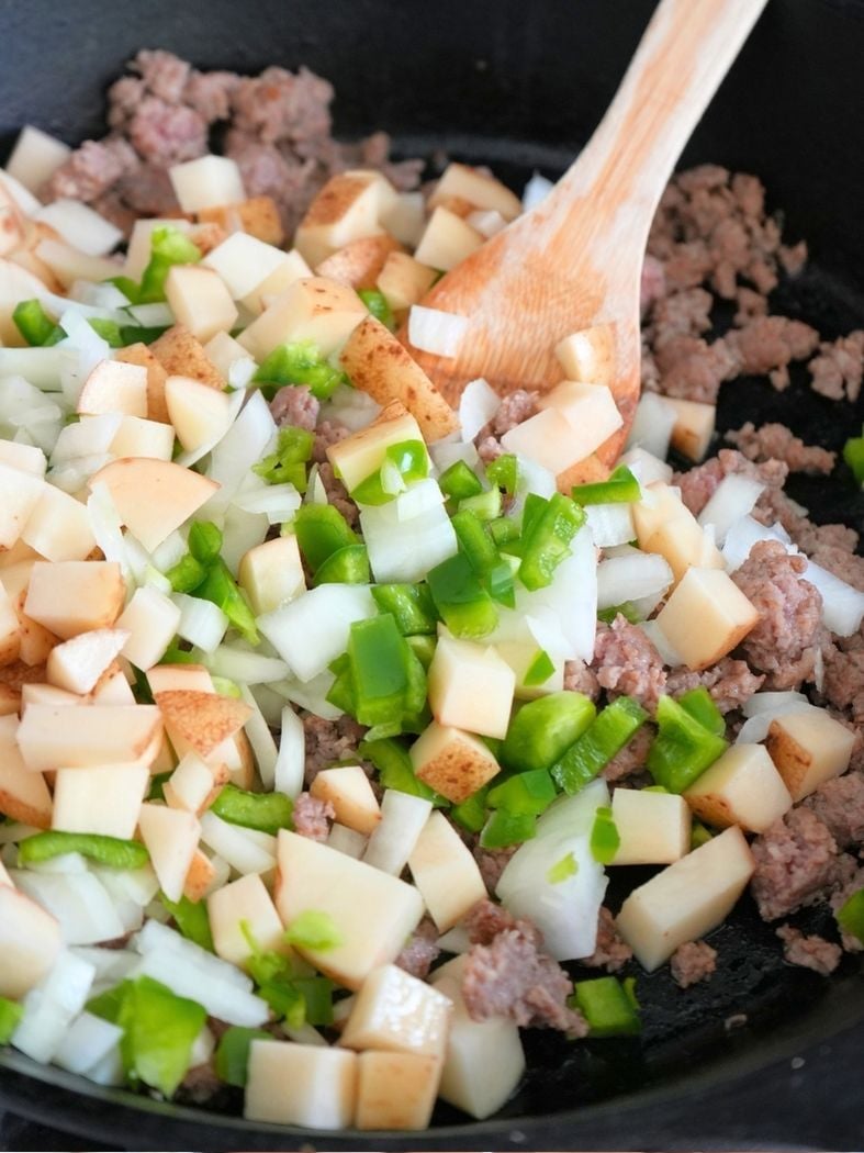 Onions, peppers, and potatoes on browned sausage in skillet, shown with wooden spoon