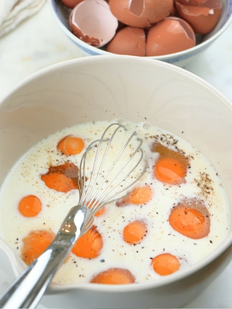 Eggs and milk in a mixing bowl, shown with a whisk in the bowl and egg shells in the background