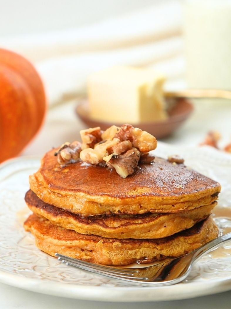 A stack of pumpkin spice pancakes topped with walnuts with butter and a pumpkin in the background