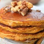A close up of a stack of pumpkin spice pancakes on a plate, topped with walnut and syrup