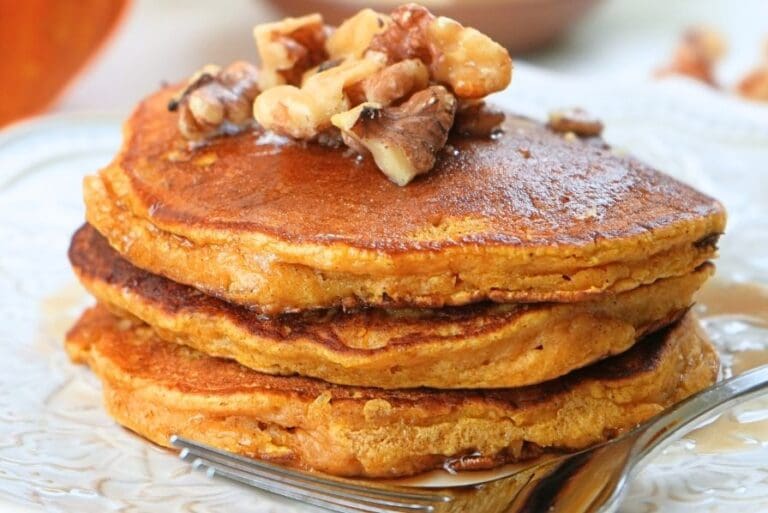 A close up of a stack of pumpkin spice pancakes on a plate, topped with walnut and syrup
