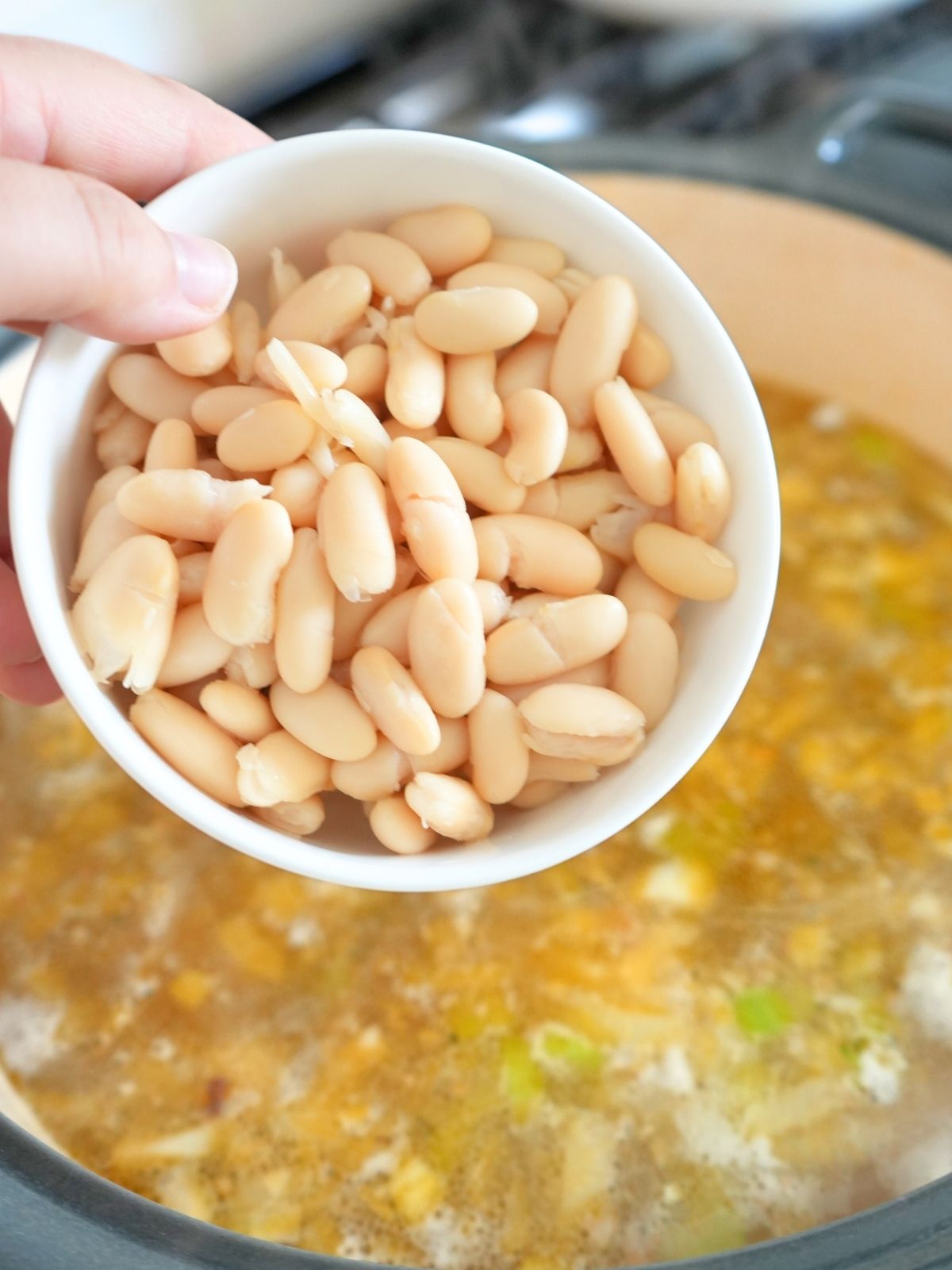 A hand holding a bowl of cannellini beans over a pot of soup.