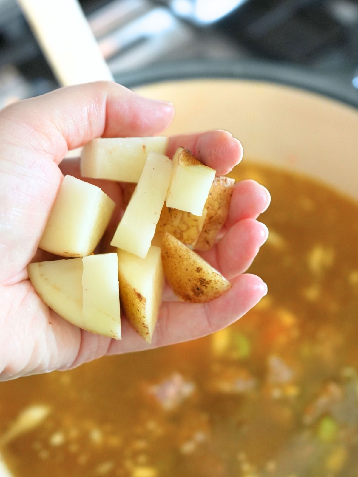A hand holding chopped potatoes over a pot of soup.