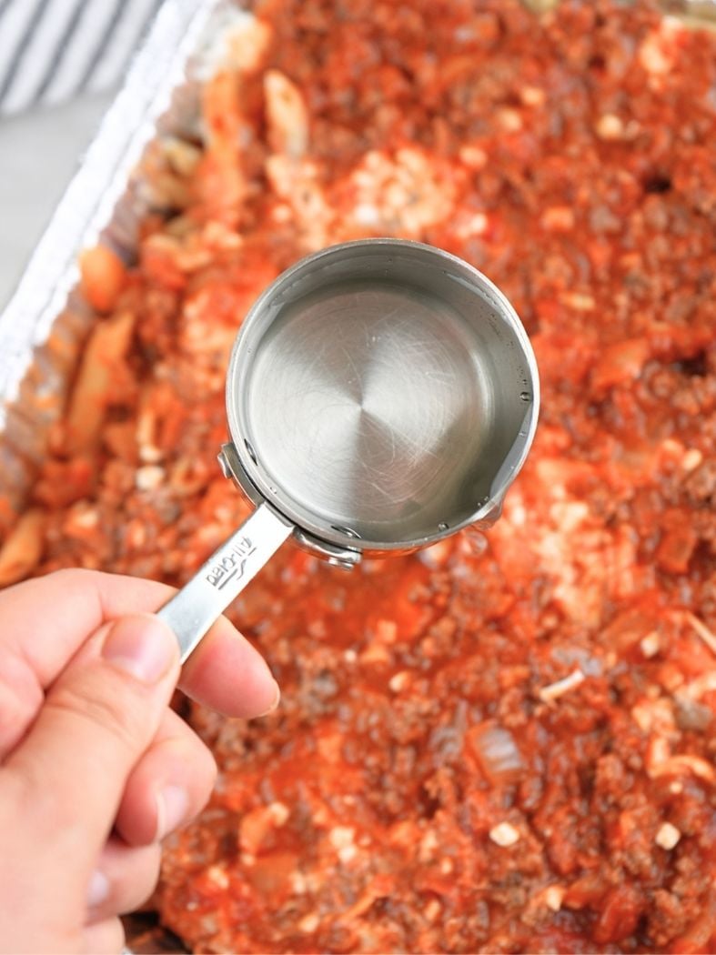 A hand holding a measuring cup filled with water over a pasta dish