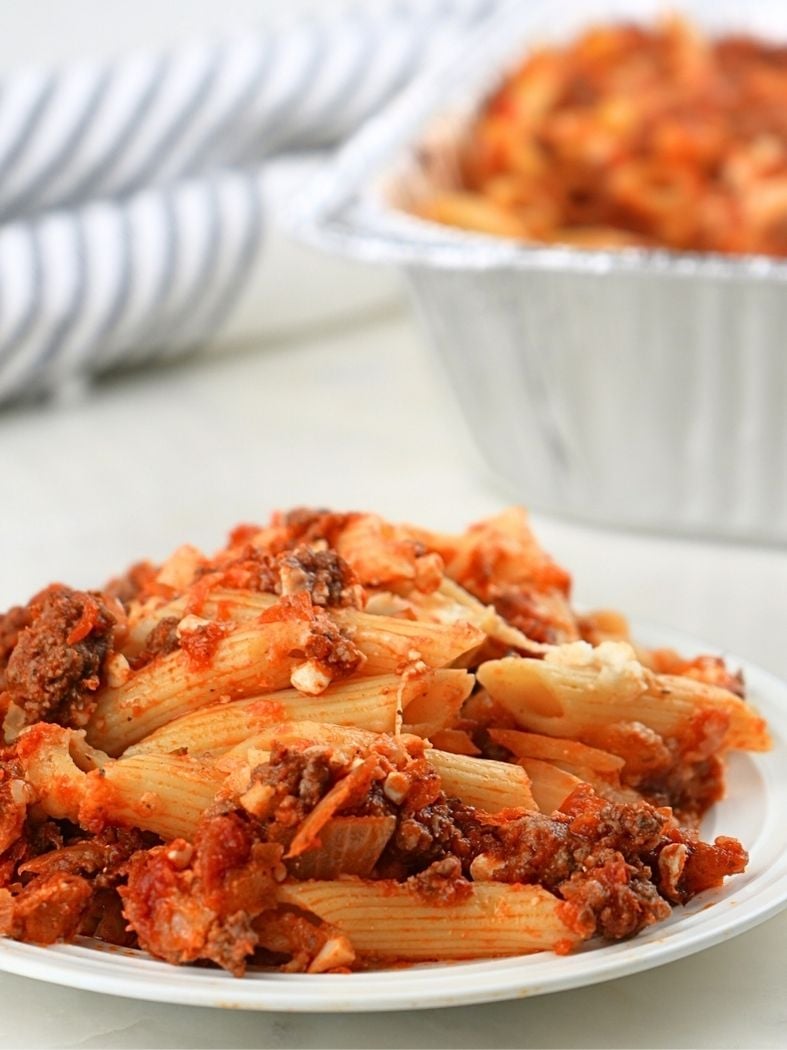 A plate of baked ziti in the foreground with the freezer meal in a foil pan in the background
