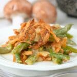 A scoop of caramelized onion green bean casserole on a plate with mushrooms and a baking dish in the background