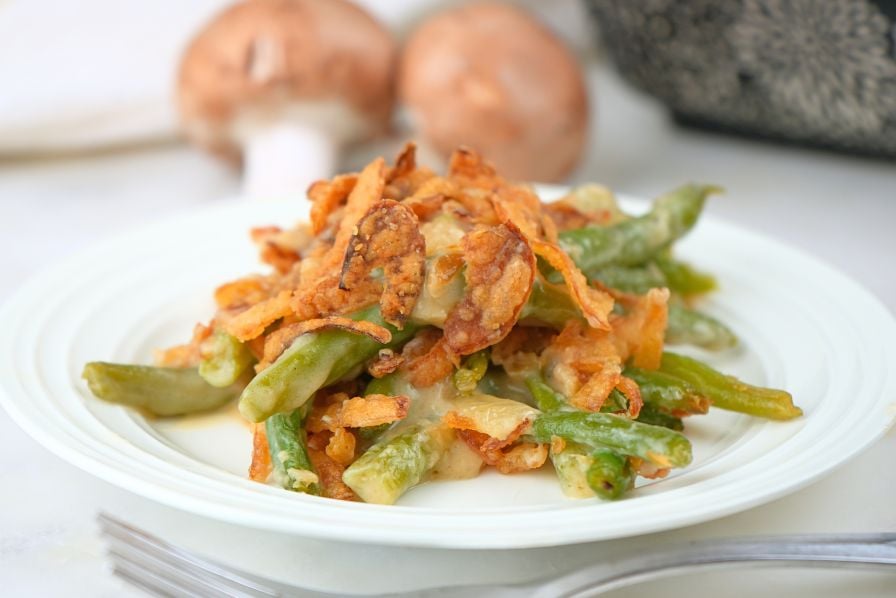 A scoop of caramelized onion green bean casserole on a plate with mushrooms and a baking dish in the background