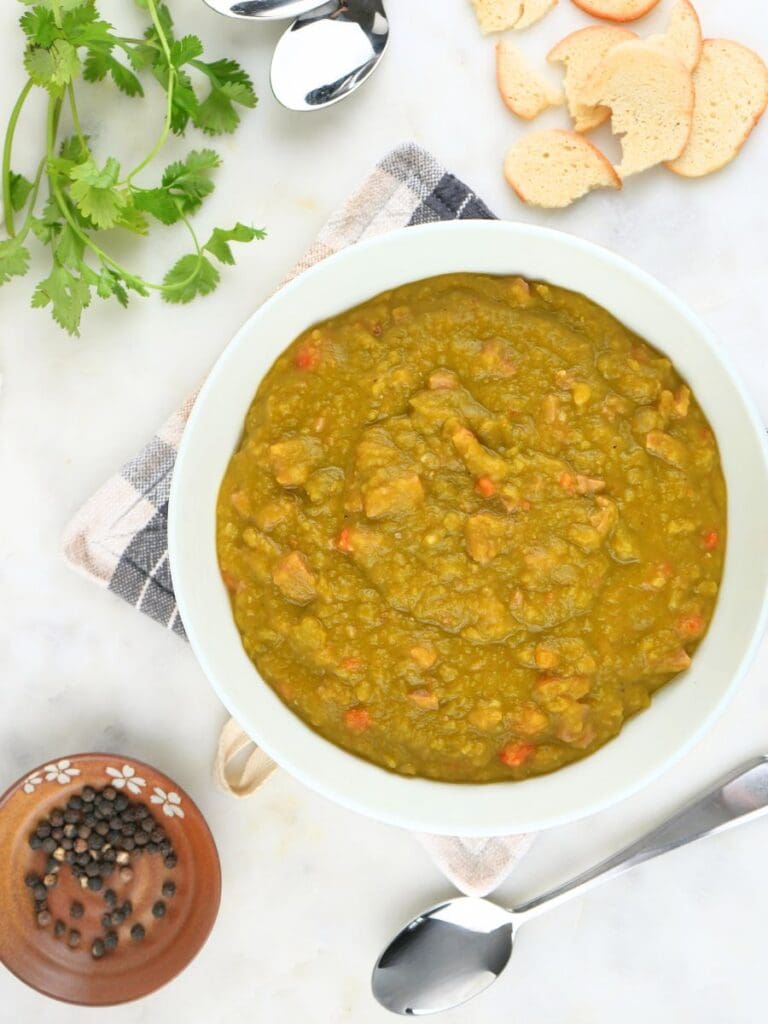 Overhead of a bowl of split pea soup with herbs, spoons, crackers, and peppercorns scattered on the counter