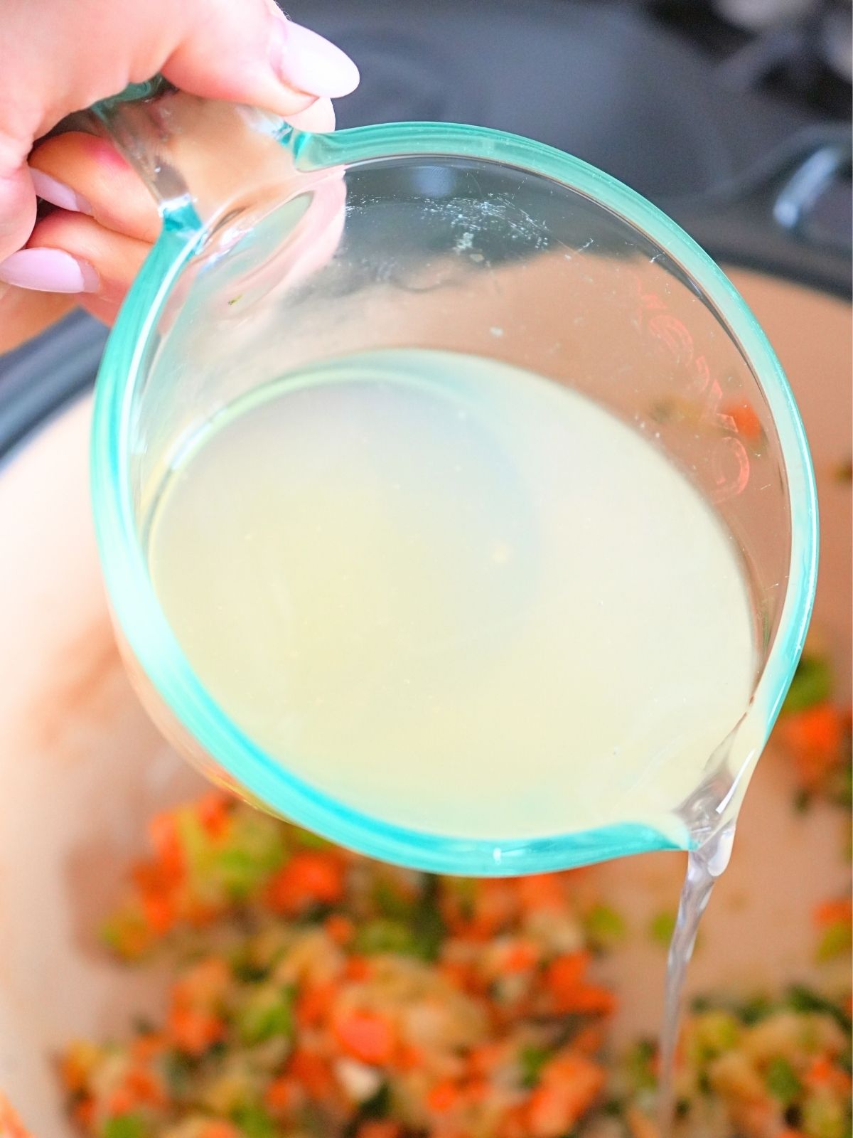 A measuring cup of chicken broth being poured into a soup pot.