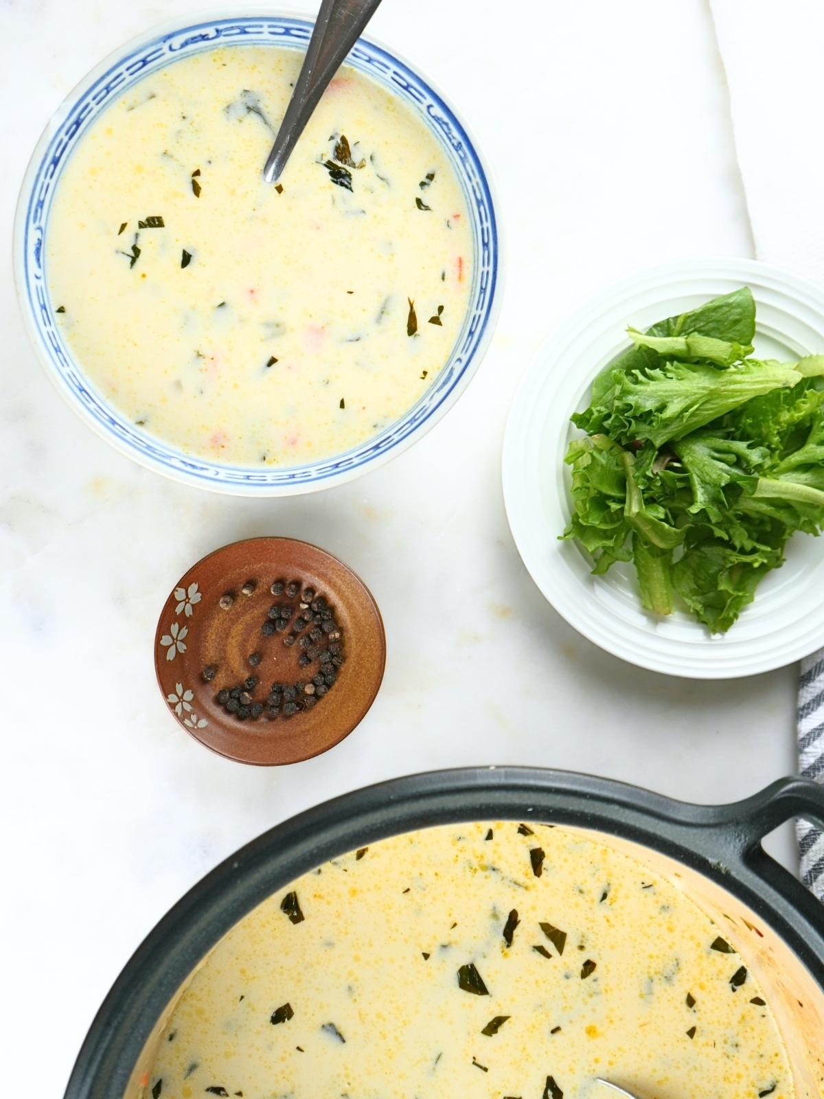 A bowl of creamy gnocchi soup next to salad, peppercorns, and a pot of soup on a counter.