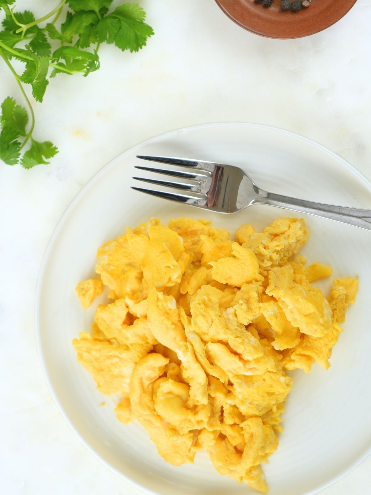 Fluffy scrambled eggs on a plate, next to fresh herbs and peppercorns.