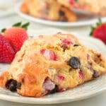 A strawberry chocolate chip sourdough scone on a plate with fresh berries scattered in the background.