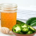 A glass jar of jalapeño hot honey with fresh peppers and a honeycomb stick.