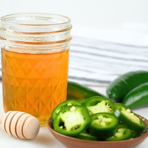 A glass jar of jalapeño hot honey with fresh peppers and a honeycomb stick.