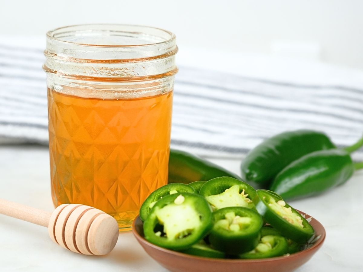 A glass jar of jalapeño hot honey with fresh peppers and a honeycomb stick.