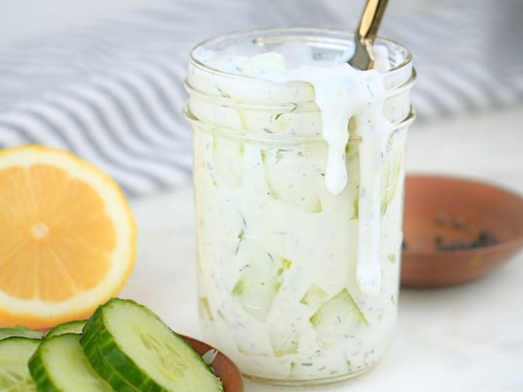 Cucumber ranch dressing in a jar with fresh cucumbers and lemons next to it.